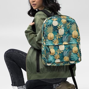 May include: A teal backpack with a repeating pattern of gold and white pineapples and green leaves. The backpack has black straps and a black zipper. The person in the image is wearing a green jacket, black jeans, and tan shoes.