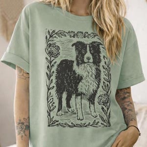 May include: A light green t-shirt featuring a black and white illustration of a dog within a floral frame. The dog has a black and white coat and is standing. The t-shirt has rolled-up sleeves.