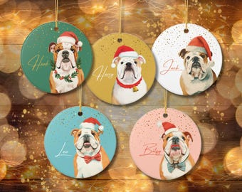 May include: Five round Christmas ornaments featuring bulldog illustrations wearing Santa hats. Each ornament has a different background color: green, gold, white, blue, and pink. Names are written on each ornament: Hank, Harry, Jack, Leo, and Betsy.