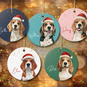 Puede incluir: Cinco adornos redondos de cerámica con ilustraciones de perros beagle que llevan gorros de Papá Noel rojos y blancos. Cada adorno muestra un color de fondo único, motas doradas y un nombre escrito en cursiva: Belle, Willow, Daisy, Elsie y Holly.
