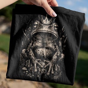 May include: A black t-shirt featuring a detailed, monochromatic graphic print of a frog wearing a crown. The frog is positioned centrally, surrounded by stylized botanical elements including ferns and small flowers, rendered in shades of off-white.