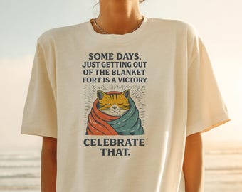 May include: Cream-colored t-shirt featuring a graphic of a cat wrapped in a blanket. The cat is orange and yellow, with a blue and red blanket. Text on the shirt reads, "SOME DAYS, JUST GETTING OUT OF THE BLANKET FORT IS A VICTORY. CELEBRATE THAT."