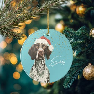 May include: Round Christmas ornament featuring a German Shorthaired Pointer wearing a Santa hat. The dog's portrait is set against a light blue background with gold confetti and the name "Eddy" in white script. A gold string is attached for hanging.