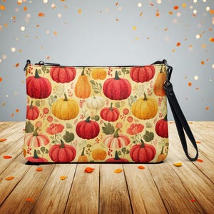 May include: A rectangular clutch with a black wrist strap. The clutch features a repeating pattern of pumpkins in various colors, including red, orange, yellow, and white, set against a cream-colored background with floral accents. The zipper is black.