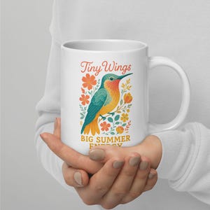 May include: White ceramic mug featuring a colorful illustration of a hummingbird with the text "Tiny Wings" and "Big Summer Energy". The hummingbird has teal, orange, and yellow feathers, and is surrounded by orange and yellow flowers.
