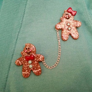 May include: Two gingerbread people ornaments connected by a silver chain. The gingerbread people are decorated with red and green glitter and have red bows.