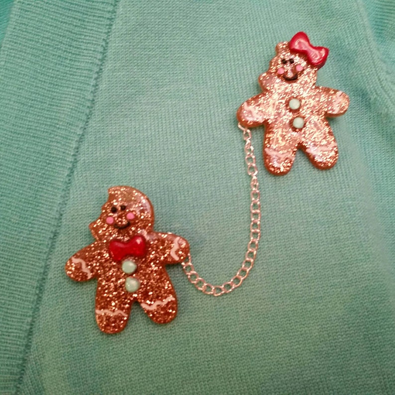 May include: Two gingerbread people ornaments connected by a silver chain. The gingerbread people are decorated with red and green glitter and have red bows.