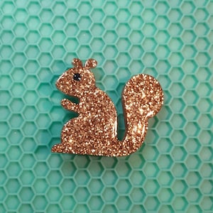 May include: A gold glitter squirrel figurine with a black eye. The squirrel is sitting on a teal honeycomb background.