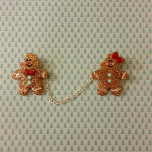 May include: Two gingerbread people ornaments connected by a silver chain. The gingerbread person on the left is wearing a red bow tie and the gingerbread person on the right is wearing a red bow. Both gingerbread people are decorated with gold glitter.