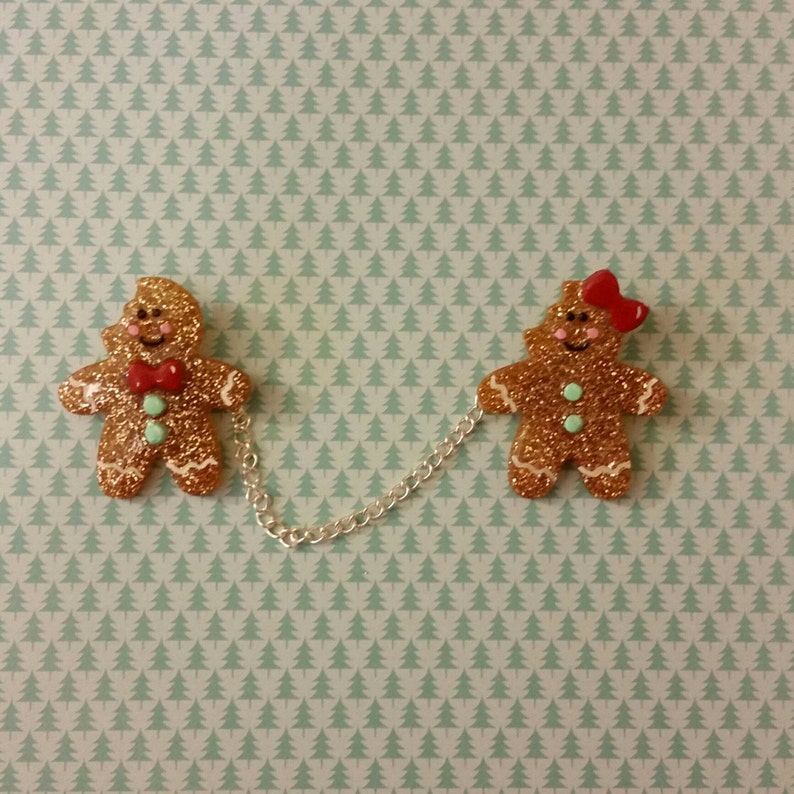 May include: Two gingerbread people ornaments connected by a silver chain. The gingerbread person on the left is wearing a red bow tie and the gingerbread person on the right is wearing a red bow. Both gingerbread people are decorated with gold glitter.
