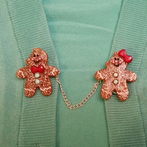May include: Two gingerbread person-shaped brooches with glitter and red accents connected by a silver chain. The brooches are on a green sweater.