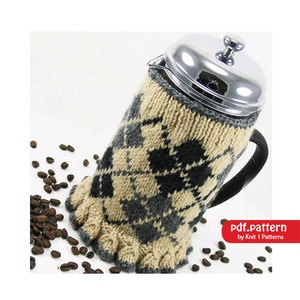 May include: A knitted cosy in a beige and grey argyle pattern with black accents fits a French press coffee pot. The cosy has a ruffled edge. Coffee beans are scattered on a white surface.