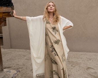 May include: A beige sleeveless maxi dress featuring a dark green botanical fern print pattern, paired with a long, loose-fitting white open-front kimono-style cardigan with fringe detailing at the hemline and brown leather boots.