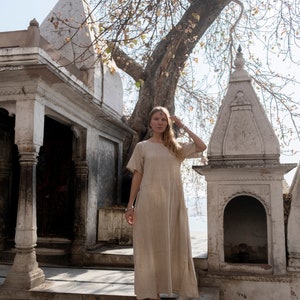 Puede incluir: Una mujer con un vestido de lino marr&oacute;n claro se encuentra en las escaleras de un templo blanco. El templo tiene un techo alto y puntiagudo y tallas intrincadas. La mujer mira a la c&aacute;mara y tiene la mano en la cabeza. El fondo es un bosque verde exuberante.
