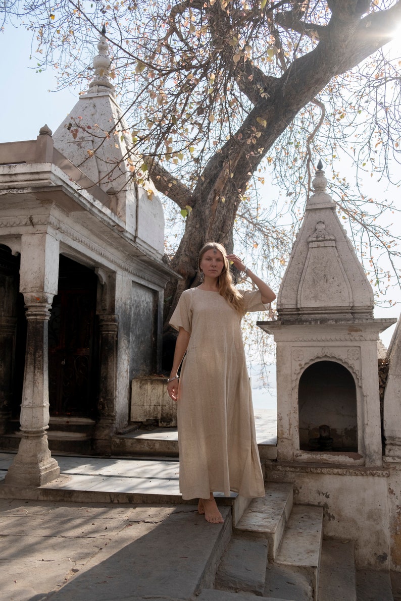 Puede incluir: Una mujer con un vestido de lino marr&oacute;n claro se encuentra en las escaleras de un templo blanco. El templo tiene un techo alto y puntiagudo y tallas intrincadas. La mujer mira a la c&aacute;mara y tiene la mano en la cabeza. El fondo es un bosque verde exuberante.