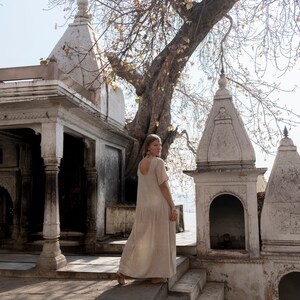 Puede incluir: Una mujer con un vestido largo de color claro camina junto a un templo blanco con detalles intrincados. El templo est&aacute; rodeado de &aacute;rboles y vegetaci&oacute;n.
