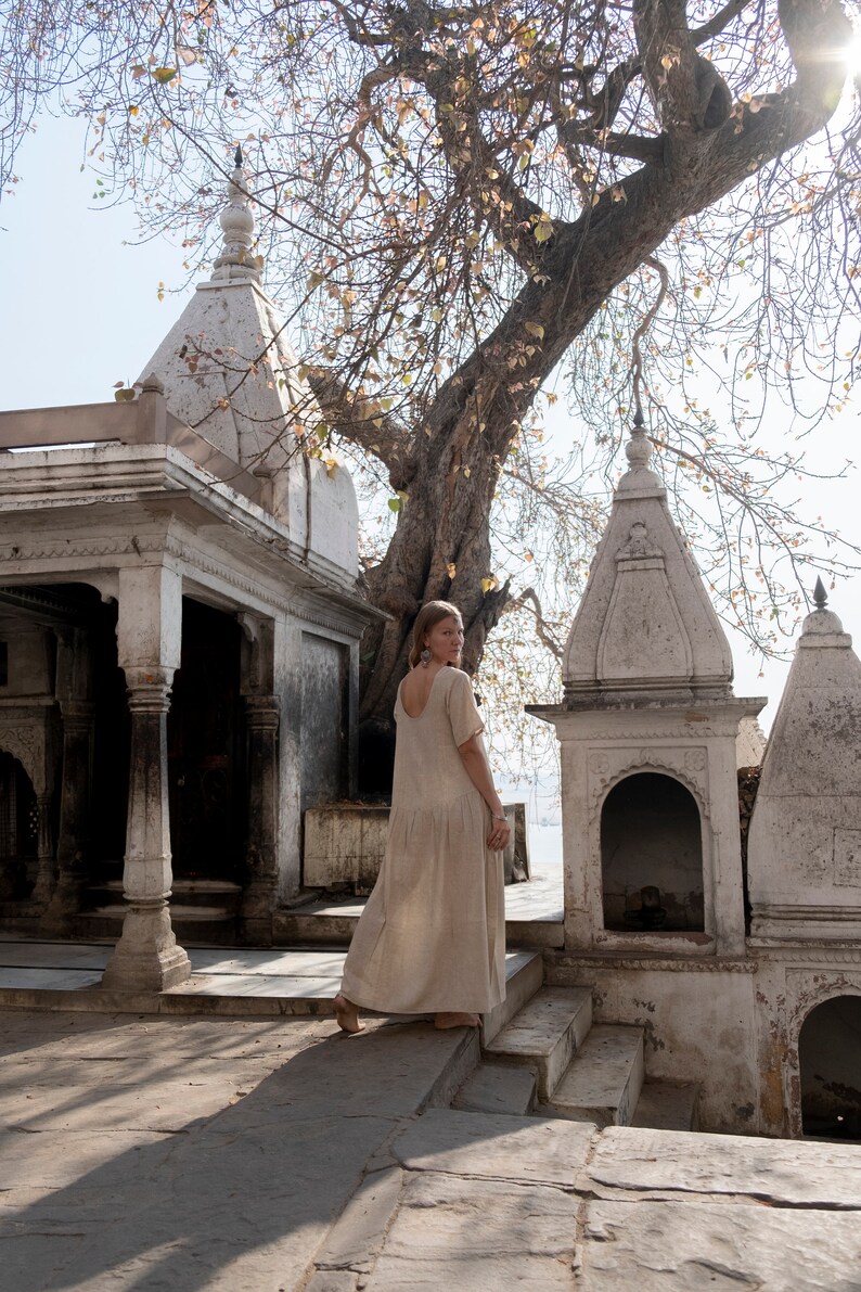 Puede incluir: Una mujer con un vestido largo de color claro camina junto a un templo blanco con detalles intrincados. El templo est&aacute; rodeado de &aacute;rboles y vegetaci&oacute;n.