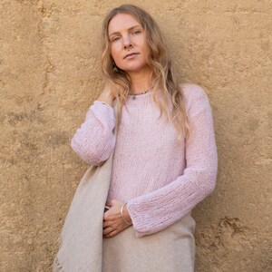 May include: A woman wearing a light pink knitted sweater and a beige shawl. She is standing in front of a textured yellow wall.
