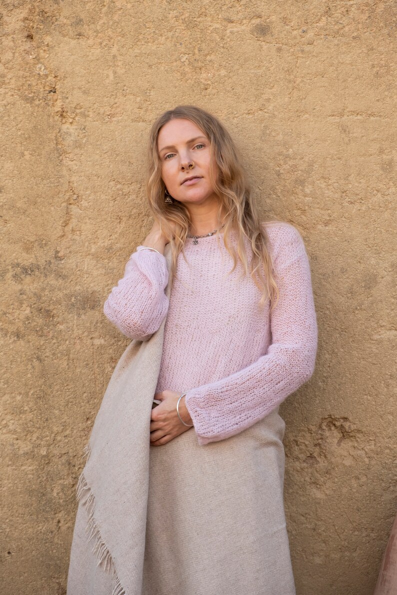 May include: A woman wearing a light pink knitted sweater and a beige shawl. She is standing in front of a textured yellow wall.