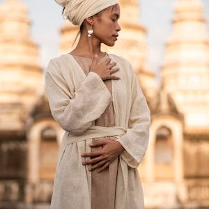 May include: A woman wearing a white linen robe with a matching head wrap and pearl earrings. She is standing in front of a large, ornate building with a golden hue.