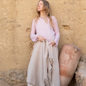 May include: A woman wearing a light pink sweater and a beige shawl stands in front of a tan wall. She is barefoot and has long blonde hair. A large terracotta pot is leaning against the wall next to her.