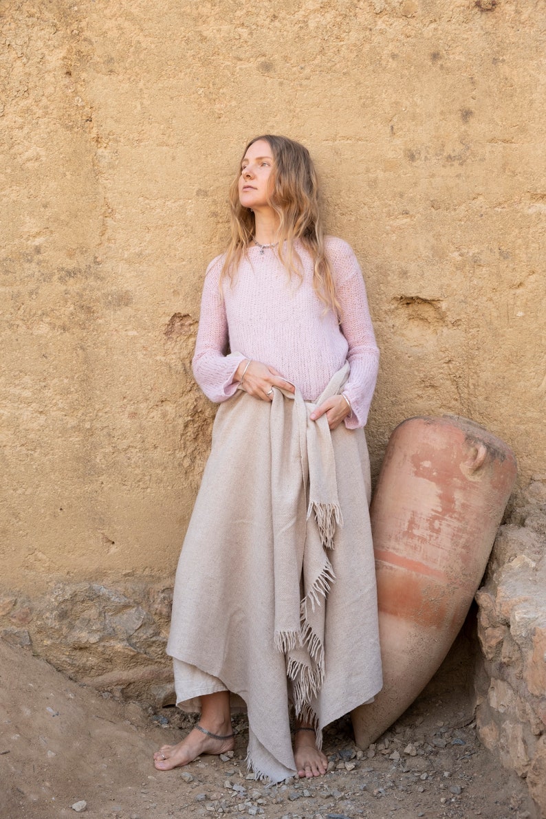 May include: A woman wearing a light pink sweater and a beige shawl stands in front of a tan wall. She is barefoot and has long blonde hair. A large terracotta pot is leaning against the wall next to her.