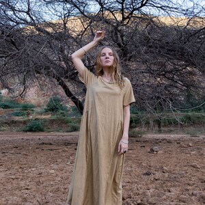 May include: A woman wearing a long, light brown, loose-fitting dress stands in a field with bare feet. The field is dry and cracked. A bare tree with branches reaching up is behind the woman.
