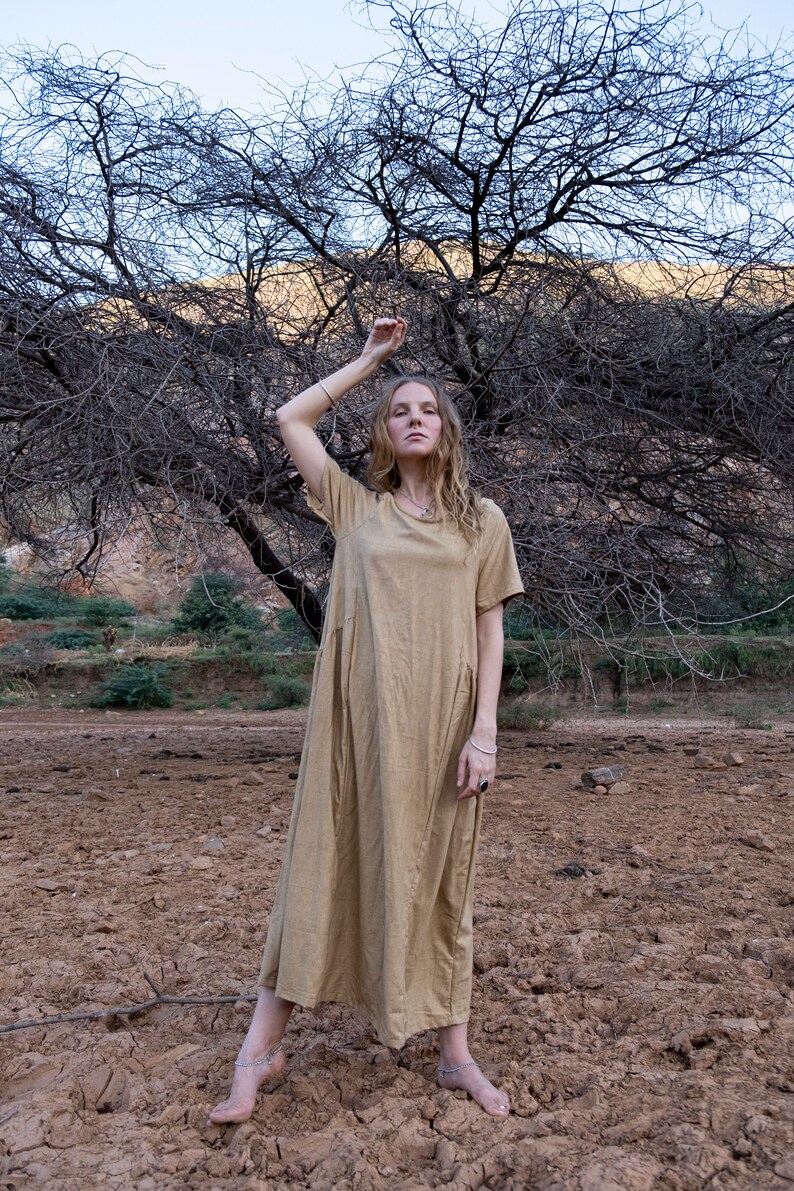May include: A woman wearing a long, light brown, loose-fitting dress stands in a field with bare feet. The field is dry and cracked. A bare tree with branches reaching up is behind the woman.