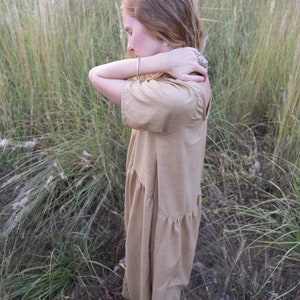 May include: A woman wearing a light brown, tiered dress stands in a field of tall grass. She is looking down and has her arms crossed in front of her.