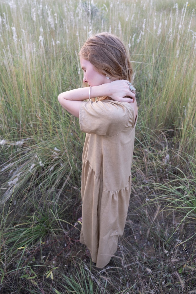 May include: A woman wearing a light brown, tiered dress stands in a field of tall grass. She is looking down and has her arms crossed in front of her.