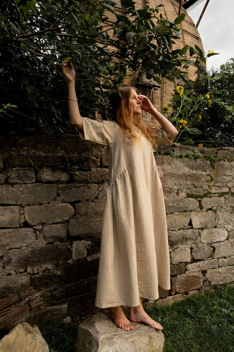 Puede incluir: Una mujer con un vestido largo de lino beige claro se para sobre una piedra frente a un muro de piedra. Ella est&aacute; descalza y tiene el pelo largo y suelto. La pared est&aacute; cubierta de musgo y enredaderas.