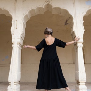 May include: A woman wearing a black dress stands in a white archway. She is wearing a headscarf and has her back to the camera.