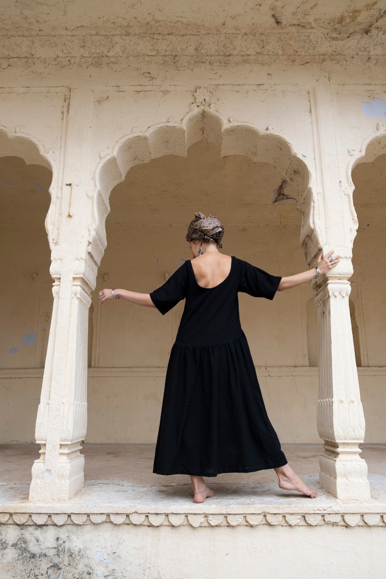 May include: A woman wearing a black dress stands in a white archway. She is wearing a headscarf and has her back to the camera.