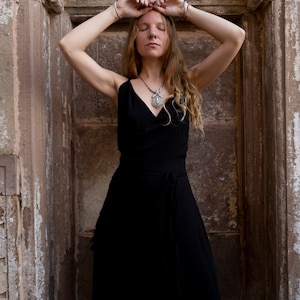 Puede incluir: Vestido negro cruzado con cuello en V y cintura anudada. El vestido se lleva con un collar con colgante plateado y pulseras. La persona está de pie frente a una pared de piedra con los brazos levantados.