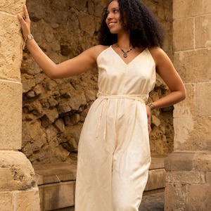 Könnte beinhalten: Cremefarbener Jumpsuit mit V-Ausschnitt und passendem Gürtel. Das Model trägt eine Halskette und ein Armband. Der Hintergrund ist eine Steinmauer. Der Jumpsuit ist aus einem weichen, fließenden Stoff.
