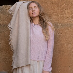 May include: A woman wearing a light pink knitted sweater, a long beige skirt, and a beige shawl draped over her shoulders. She is standing in front of a textured brown wall.
