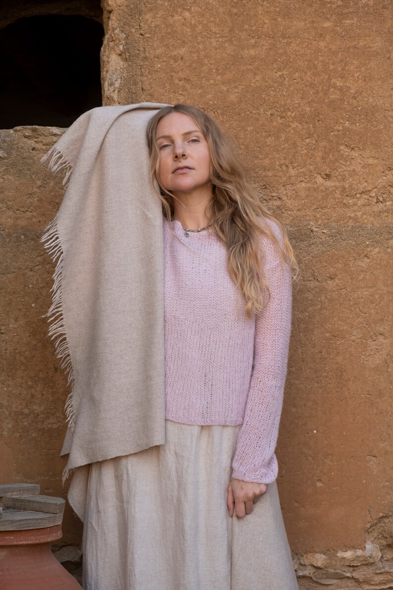 May include: A woman wearing a light pink knitted sweater, a long beige skirt, and a beige shawl draped over her shoulders. She is standing in front of a textured brown wall.