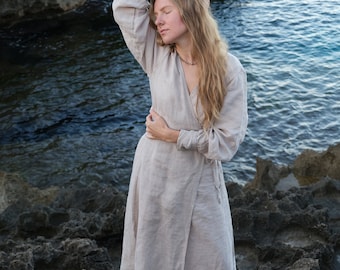 May include: A woman wearing a light beige linen dress stands on a rocky shore with the ocean in the background. She is looking down and has her hand on her head.