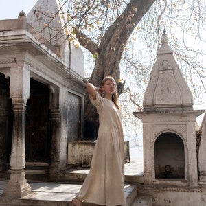 Puede incluir: Una mujer con un vestido de lino beige se encuentra en un conjunto de escalones de piedra frente a un templo blanco con un &aacute;rbol en el fondo.