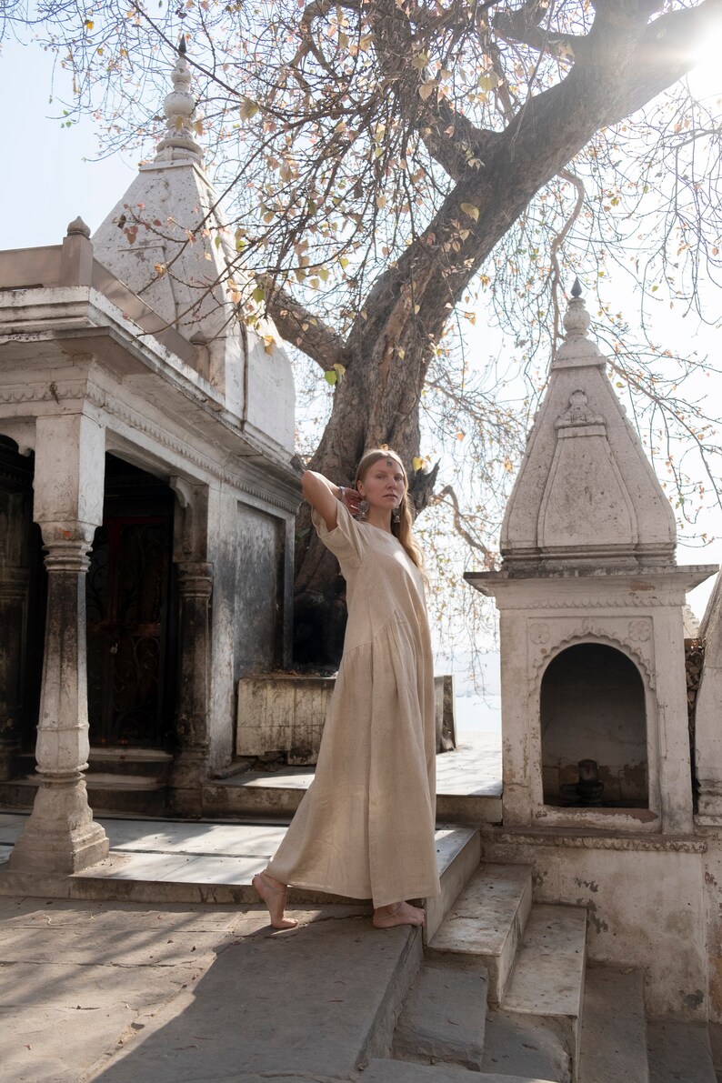 Puede incluir: Una mujer con un vestido de lino beige se encuentra en un conjunto de escalones de piedra frente a un templo blanco con un &aacute;rbol en el fondo.