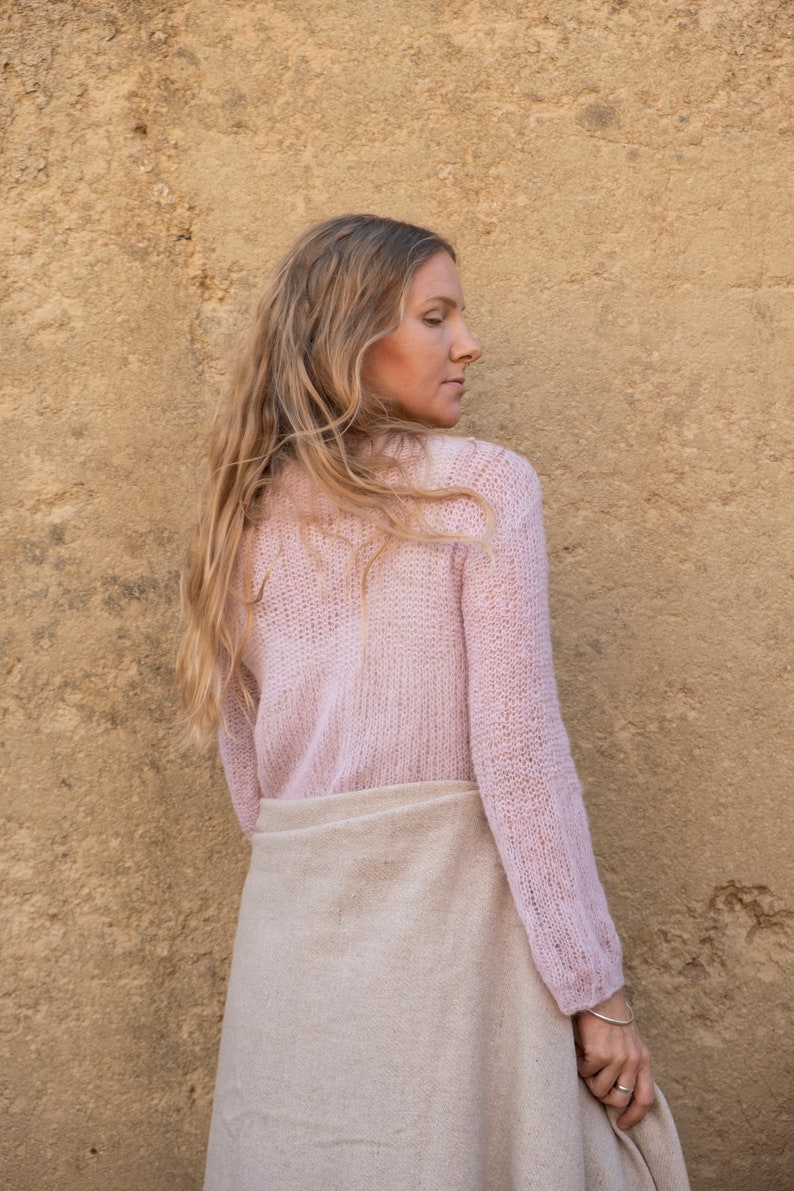 May include: A woman wearing a light pink knitted sweater and a beige linen skirt. She is standing in front of a textured yellow wall.