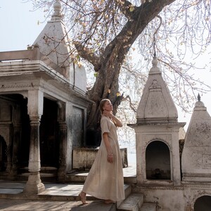 Puede incluir: Una mujer con un vestido marr&oacute;n claro se encuentra frente a un templo de piedra blanca con un gran &aacute;rbol detr&aacute;s de ella. El templo tiene tallas intrincadas y un techo alto y puntiagudo.