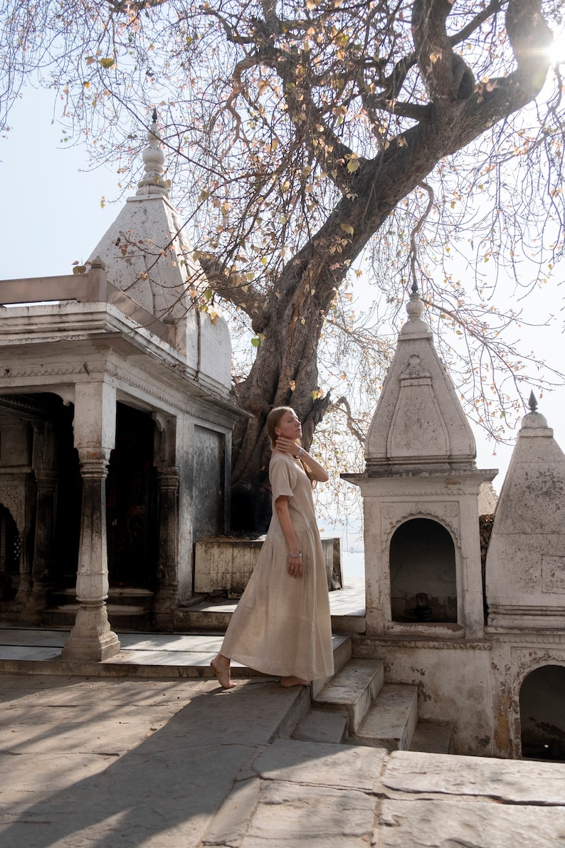 Puede incluir: Una mujer con un vestido marr&oacute;n claro se encuentra frente a un templo de piedra blanca con un gran &aacute;rbol detr&aacute;s de ella. El templo tiene tallas intrincadas y un techo alto y puntiagudo.