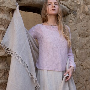 May include: A woman wearing a light pink knitted sweater with a light brown shawl draped around her shoulders. She is standing in front of a stone wall.