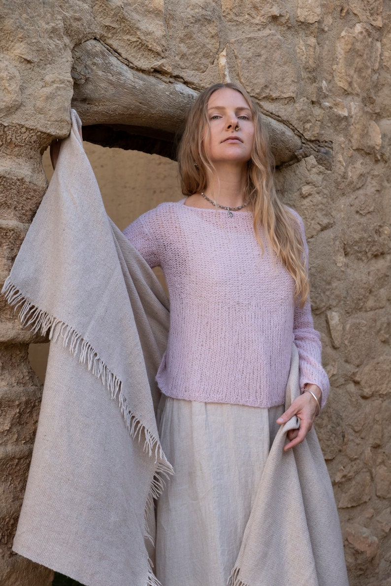 May include: A woman wearing a light pink knitted sweater with a light brown shawl draped around her shoulders. She is standing in front of a stone wall.