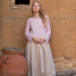 May include: A woman wearing a light pink sweater and a long, beige skirt stands in front of a rustic stone wall. She is barefoot and holding the skirt in front of her. A large terracotta pot is to the left of the woman.