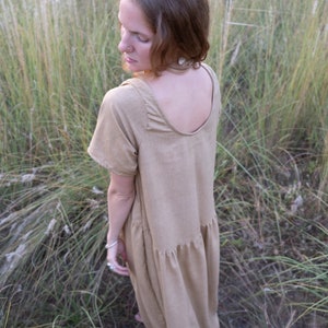 May include: A woman wearing a light brown, loose-fitting, knee-length dress with a scoop neck and short sleeves. She is standing in a field of tall grass.