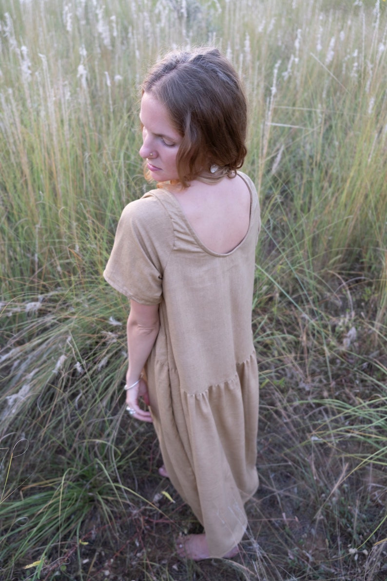May include: A woman wearing a light brown, loose-fitting, knee-length dress with a scoop neck and short sleeves. She is standing in a field of tall grass.