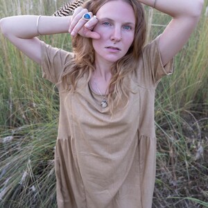 May include: A woman wearing a light brown dress with a silver necklace and a silver ring with a blue stone. She is standing in a field of tall grass.