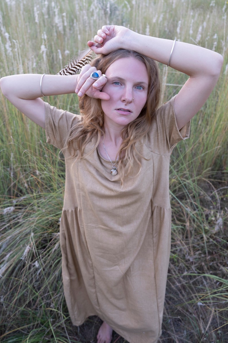 May include: A woman wearing a light brown dress with a silver necklace and a silver ring with a blue stone. She is standing in a field of tall grass.
